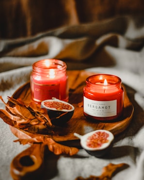 Two lit red scented candles labeled Cinnamon and Bergamot are placed on a wooden tray. Accompanying the candles are dried autumn leaves and halved figs, all set against a soft fabric background.