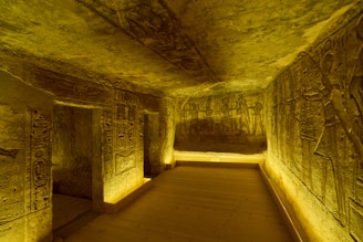 The image depicts an ancient Egyptian tomb with walls intricately carved with hieroglyphics and figures. The space is illuminated with a warm, golden light that highlights the detailed engravings. The room is narrow with a wooden floor and stone walls, and the carvings appear to tell a story or display scenes from ancient Egyptian mythology.