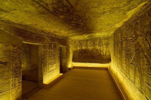 The image depicts an ancient Egyptian tomb with walls intricately carved with hieroglyphics and figures. The space is illuminated with a warm, golden light that highlights the detailed engravings. The room is narrow with a wooden floor and stone walls, and the carvings appear to tell a story or display scenes from ancient Egyptian mythology.
