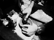 A pair of hands is grinding coffee beans in a mortar with a pestle. The scene includes a moka pot beside the grinder, indicating a traditional coffee-making process. Sunlight creates strong shadows over a woven mat on the ground, adding texture.
