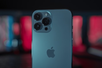 A modern smartphone with a sleek silver finish is prominently featured, showcasing its advanced triple camera setup. The background is softly blurred with contrasting red and blue hues, giving it a minimalist and stylish appearance.