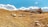 A group of sheep grazes on a sunlit, grassy field with gentle rolling hills. An old barn with a slanted roof is situated on the right, slightly elevated. The sky is bright blue with scattered fluffy clouds.