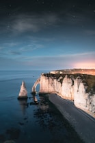 A picturesque seascape featuring dramatic limestone cliffs jutting out into the ocean. The sky is a deep twilight hue speckled with stars, creating a serene and tranquil atmosphere. The cliffs are illuminated with soft, warm lighting, highlighting their rugged texture.