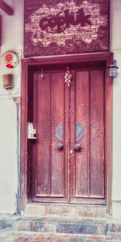 A rustic wooden double door with decorative circular metal elements at the center is set into a brick and plaster facade. Above the door, there is a brick sign with text. An old-fashioned lantern hangs on the right side, and there is a small decorative charm attached to the door. A security alarm and an intercom system are mounted on the left side.