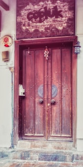 A rustic wooden double door with decorative circular metal elements at the center is set into a brick and plaster facade. Above the door, there is a brick sign with text. An old-fashioned lantern hangs on the right side, and there is a small decorative charm attached to the door. A security alarm and an intercom system are mounted on the left side.