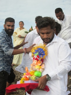 Several people are gathered outdoors, with one individual prominently holding a colorful statue of a Hindu deity. The statue is adorned with decorative items, including a golden headdress and various trinkets. The group appears to be engaged in a ceremonial or religious activity, with some individuals looking on attentively.