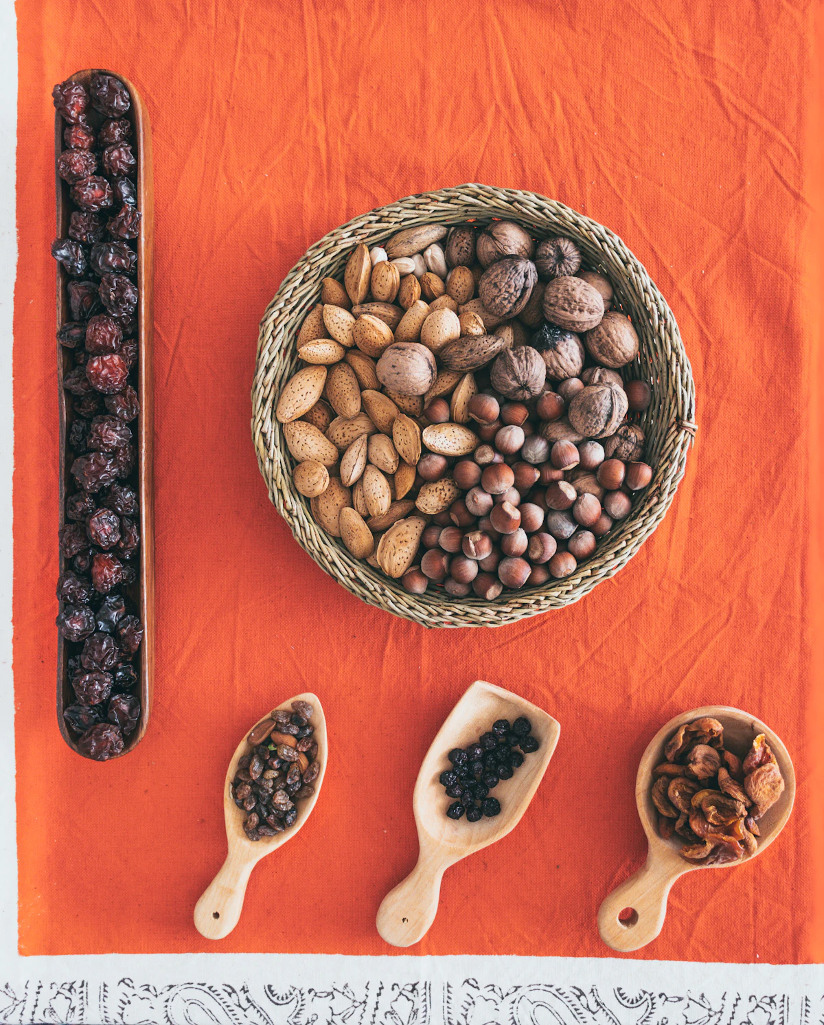 A woven tray of mixed nuts with wooden spoons of raisins and dried berries on a bright cloth