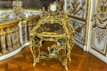 An ornate, antique desk with intricate gilt detailing and a small decorative clock on top. The surrounding decor includes lavish gold accents and detailed molding on the walls, reflecting a luxurious and historical style.