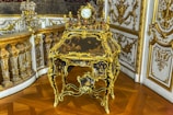 An ornate, antique desk with intricate gilt detailing and a small decorative clock on top. The surrounding decor includes lavish gold accents and detailed molding on the walls, reflecting a luxurious and historical style.