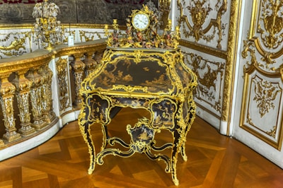 An ornate, antique desk with intricate gilt detailing and a small decorative clock on top. The surrounding decor includes lavish gold accents and detailed molding on the walls, reflecting a luxurious and historical style.