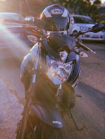 A close-up view of a Yamaha motorcycle parked outdoors with visible lens flare effects from the sun. The motorcycle is sleek with a black and silver design, and a helmet is placed on the seat. Several cars are visible in the background.