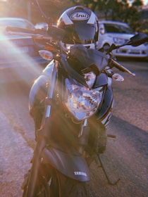 A close-up view of a Yamaha motorcycle parked outdoors with visible lens flare effects from the sun. The motorcycle is sleek with a black and silver design, and a helmet is placed on the seat. Several cars are visible in the background.