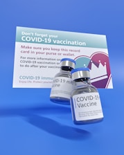 Two vials labeled 'COVID-19 Vaccine' are prominently placed in front of a vaccination reminder card. The card emphasizes keeping the record in a purse or wallet and has informational text about the vaccine. The background is a solid blue color.