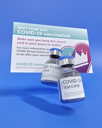 Two vials labeled 'COVID-19 Vaccine' are prominently placed in front of a vaccination reminder card. The card emphasizes keeping the record in a purse or wallet and has informational text about the vaccine. The background is a solid blue color.