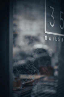 A blurred reflection of a street scene is visible through a glass window. The number 35 and partial lettering indicating a business sign are present. The background is out of focus, featuring hints of a dark interior with plant outlines and possible furnishings.