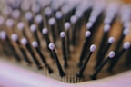 Close-up of a hairbrush with black bristles tipped with light purple rounded ends. The image is focused on the bristles, creating a pattern across the frame, with a soft background blur.