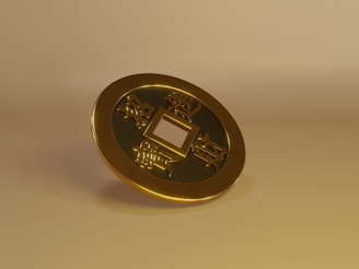 A three-dimensional rendering of a gold Chinese coin with a square hole in the center. The coin features traditional Chinese characters engraved on its surface. It is placed against a neutral, gold-toned background, enhancing its metallic sheen.