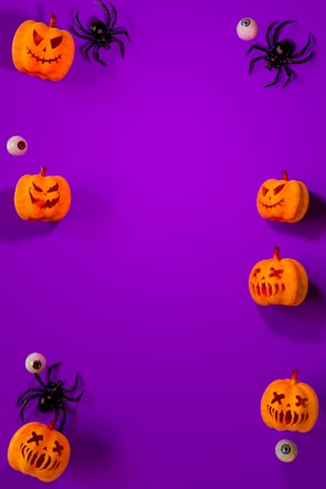 A vibrant purple background is adorned with scattered Halloween-themed items along the edges, including carved pumpkins with various facial expressions, artificial spiders, and plastic eyeballs, creating a playful and spooky arrangement.
