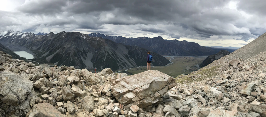 A lone trekker pausing on a rocky ridge overlooking a misty mountain valley at dawn.