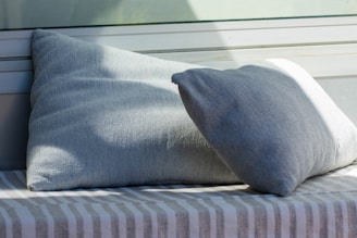 Two light gray cushions are placed on a surface covered with a gray and white striped fabric. The background features a partial view of a window and some sunlight creating shadows on the cushions.
