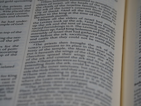 A close-up view of an open book with black text on white pages. The text is aligned in columns and features a narrative style, likely from a religious or historical context.