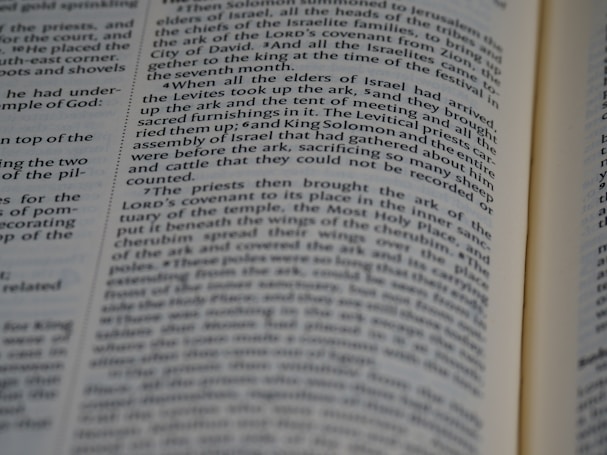 A close-up view of an open book with black text on white pages. The text is aligned in columns and features a narrative style, likely from a religious or historical context.