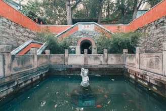 In front of an ancient structure with stone walls and red accents, there's a small pond featuring a central sculpture of a figure. Surrounding this water are stone railings and lush greenery. Several colorful fish swim in the pond, adding life to the serene scene.