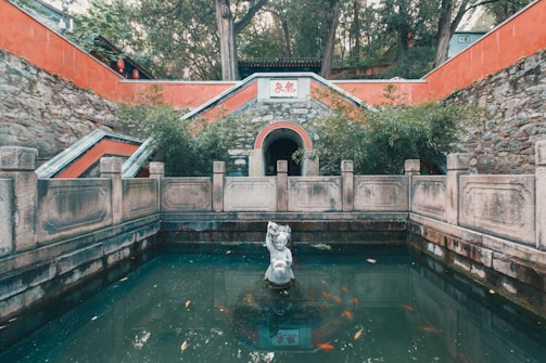 In front of an ancient structure with stone walls and red accents, there's a small pond featuring a central sculpture of a figure. Surrounding this water are stone railings and lush greenery. Several colorful fish swim in the pond, adding life to the serene scene.