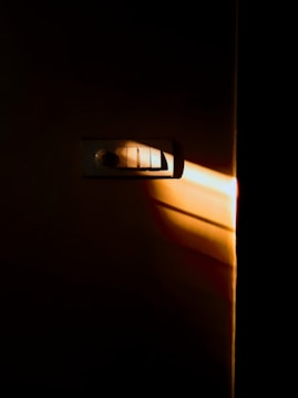 A dimly lit electrical switchboard on a wall with a beam of warm sunlight casting shadows across it. The surrounding area is dark, emphasizing the contrast between light and shadow.