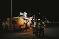 A cozy nighttime scene featuring a retro-style food truck with the sign 'Volume Cafe' illuminated against the dark sky. Warm string lights hang above, creating a welcoming ambiance. Several people are gathered around, engaging in conversation and enjoying the setting.