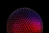 A large geodesic sphere illuminated with vibrant colors against a dark background. The structure is composed of triangular panels, radiating shades of purple and red, with small glowing points of light creating a starry effect.