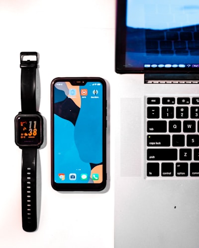 A neatly arranged set of gadgets including a black smartwatch displaying the time, a smartphone with a colorful abstract wallpaper, and a laptop with part of the keyboard visible. The devices are placed on a light surface, emphasizing their sleek design and modern technology.