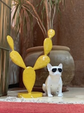 A ceramic dog wearing sunglasses is placed next to a yellow cactus sculpture, both positioned on a tiled floor. Behind them is a large terracotta-style planter containing a leafy plant. A red rug partially covers the floor in the foreground.