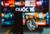 A blurred background displays a colorful television screen with various channels or streaming apps, featuring the Vietnamese word 'Quốc Tế'. In the foreground, a hand holds a smartphone on the left and a pair of wireless earphones in a case on the right, both out of focus.