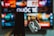 A blurred background displays a colorful television screen with various channels or streaming apps, featuring the Vietnamese word 'Quốc Tế'. In the foreground, a hand holds a smartphone on the left and a pair of wireless earphones in a case on the right, both out of focus.