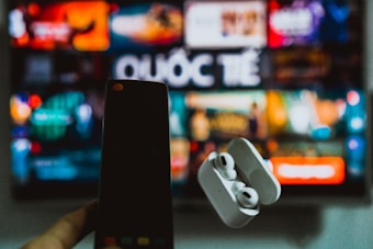 A blurred background displays a colorful television screen with various channels or streaming apps, featuring the Vietnamese word 'Quốc Tế'. In the foreground, a hand holds a smartphone on the left and a pair of wireless earphones in a case on the right, both out of focus.