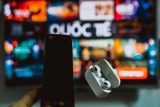 A blurred background displays a colorful television screen with various channels or streaming apps, featuring the Vietnamese word 'Quốc Tế'. In the foreground, a hand holds a smartphone on the left and a pair of wireless earphones in a case on the right, both out of focus.