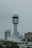 A tall, cylindrical control tower stands prominently against a cloudy sky, surrounded by modern urban buildings with a mix of architectural styles. The scene includes trees and rooftops in the foreground.