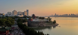 A scenic riverside landscape at sunset featuring a blend of traditional and modern architecture. In the foreground, a historic pagoda surrounded by lush greenery contrasts with the background of high-rise city buildings. People are strolling along a riverside promenade, enjoying the serene environment. The water reflects the golden hues of the sky, creating a tranquil atmosphere.