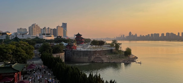 A scenic riverside landscape at sunset featuring a blend of traditional and modern architecture. In the foreground, a historic pagoda surrounded by lush greenery contrasts with the background of high-rise city buildings. People are strolling along a riverside promenade, enjoying the serene environment. The water reflects the golden hues of the sky, creating a tranquil atmosphere.