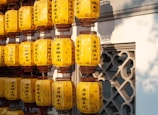 A collection of yellow paper lanterns with dark script hanging in rows against a traditional architectural backdrop. The lanterns appear to be illuminated by natural light, creating warm shadows on the intricately designed wall behind them.