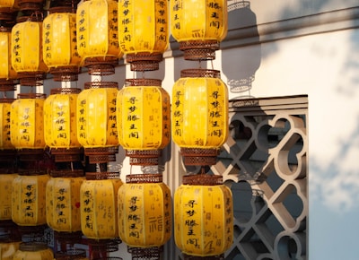 A collection of yellow paper lanterns with dark script hanging in rows against a traditional architectural backdrop. The lanterns appear to be illuminated by natural light, creating warm shadows on the intricately designed wall behind them.