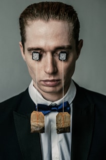 A person dressed in formal attire with tea bags hanging from their eyes. The strings of the tea bags are attached to the person's eyes using tags with QR codes. The person is wearing a dark suit, white shirt, and a blue bow tie, and has a calm expression.