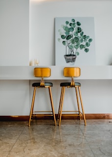 Two gold and black bar stools are positioned against a white counter with a minimalist design. Above the counter hangs a large canvas depicting a watercolor painting of a potted plant with round green leaves. The floor is a polished light brown marble, complementing the warm tones of the stools.