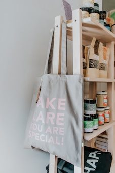 A light-grey tote bag with the words 'TAKE CARE YOU ARE SPECIAL!' printed in pink is hanging on a wooden shelf. The shelf is stocked with various jars, bottles, and bags, likely containing different food or wellness products.