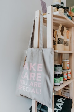 A light-grey tote bag with the words 'TAKE CARE YOU ARE SPECIAL!' printed in pink is hanging on a wooden shelf. The shelf is stocked with various jars, bottles, and bags, likely containing different food or wellness products.