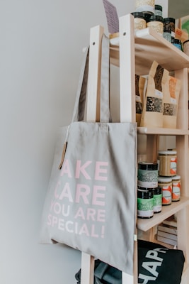 A light-grey tote bag with the words 'TAKE CARE YOU ARE SPECIAL!' printed in pink is hanging on a wooden shelf. The shelf is stocked with various jars, bottles, and bags, likely containing different food or wellness products.