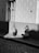 A black and white image of a city street features a woman crouching on a cobblestone sidewalk while taking a photograph of a young girl in a patterned coat. The girl stands with her back to the camera near a building wall and a window. The scene is illuminated by a contrasting shadow and light pattern.