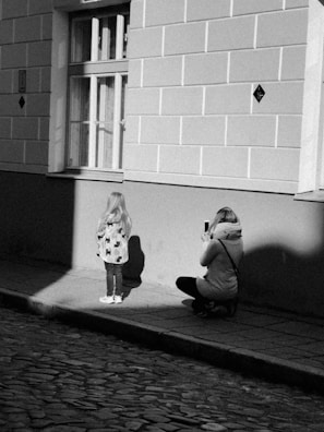 A black and white image of a city street features a woman crouching on a cobblestone sidewalk while taking a photograph of a young girl in a patterned coat. The girl stands with her back to the camera near a building wall and a window. The scene is illuminated by a contrasting shadow and light pattern.