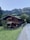 A rustic wooden cabin is nestled in a lush green landscape with mountainous scenery in the background. The cabin, with its dark wooden exterior and visible balconies, is surrounded by green trees and grass. A narrow road runs alongside the cabin, leading toward the mountains.